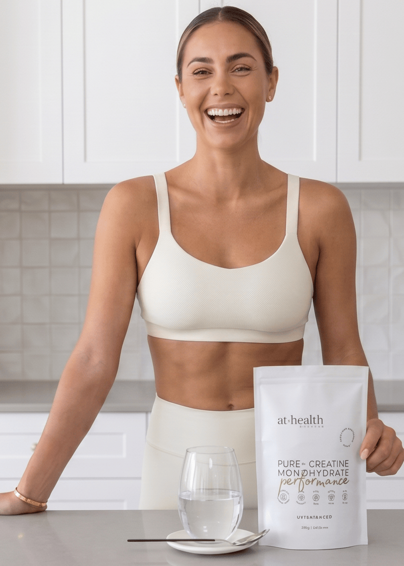 A woman in a white sports bra and matching shorts stands in a bright kitchen holding a white pouch of at:health Pure-Creatine Monohydrate Powder for Performance, with a glass of water on the counter.